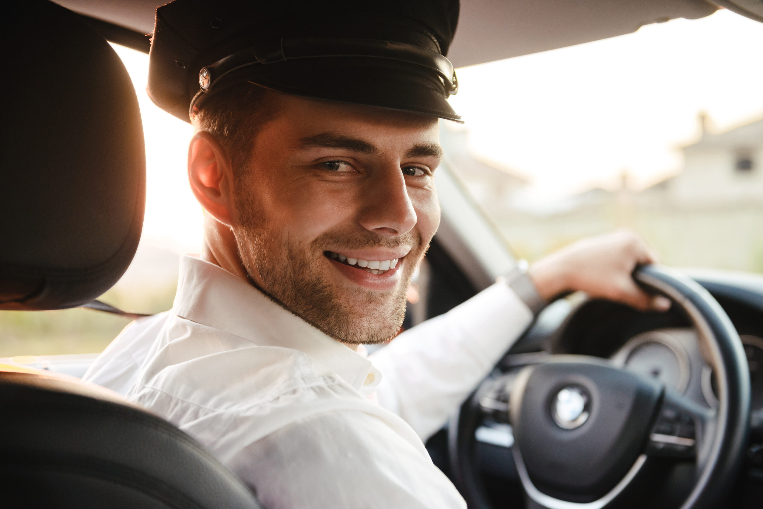 Portrait of handsome caucasian male taxi driver wearing uniform and cap smiling and looking at you while driving car